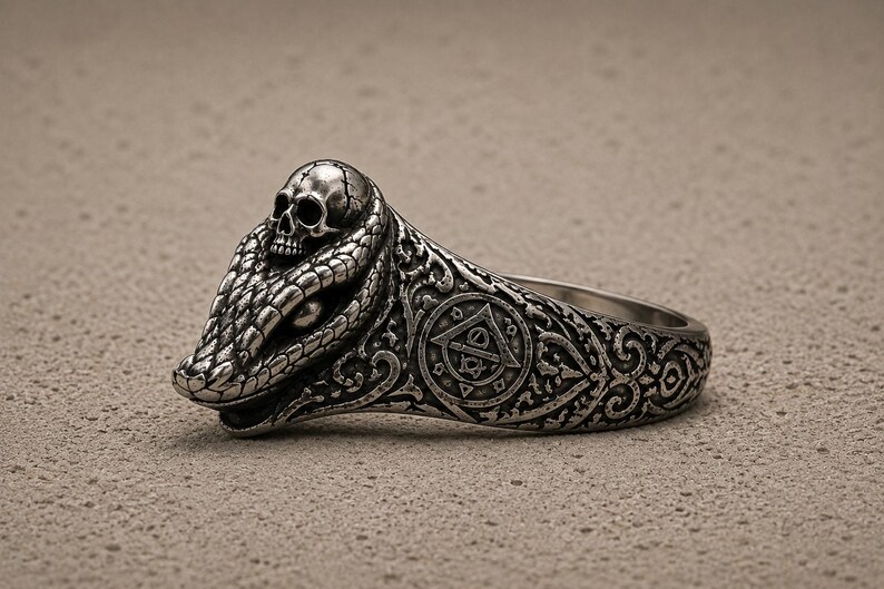 May include: A silver-toned metal ring featuring a detailed snake head with a small skull resting on top. The band is decorated with intricate scrollwork and a circular emblem containing a triangle and geometric symbols on the side.