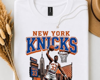 May include: White t-shirt featuring a vintage-style graphic of a basketball player dunking. The design includes the words "NEW YORK KNICKS" in orange and blue, along with an image of the Empire State Building and a basketball hoop.