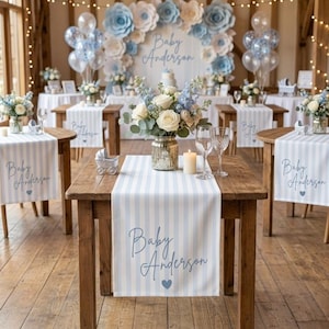 May include: A wooden table featuring a light blue and white striped runner with the text Baby Anderson and a heart icon. A glass vase holds white roses and blue flowers, accompanied by a candle, two wine glasses, and a silver carriage ornament.