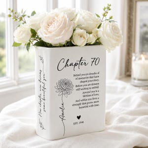 May include: A white rectangular ceramic vase filled with white roses. The front features the text Chapter 70, a floral illustration, the name Amelia, a heart icon, EST. 1946, and a poem. The side displays the text New chapter, new dreams, same beautiful you.
