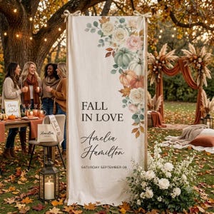 May include: A vertical fabric banner featuring a floral and pumpkin illustration in the upper right corner, with the text FALL IN LOVE, Amelia Hamilton, and SATURDAY SEPTEMBER 08 printed in black, suspended from a wooden frame with ribbon ties.