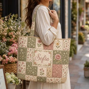 Op de afbeelding: Een gewatteerde tote bag met een patchworkontwerp van bloemenpatronen in saliegroen, oudroze en crèmetinten. Het middenpaneel toont de naam Sofia in elegant geborduurd schrift. De tas heeft twee bijpassende schouderbanden van stof.