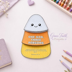 Puede incluir: Una manualidad de papel en capas con forma de caramelo de maíz con una cara sonriente en la sección blanca superior. La sección naranja central dice ONE GOD, THREE PERSONS. La sección amarilla inferior presenta el texto Baptizing them in the name of the Father and of the Son and of the Holy Spirit. Matthew 28:19.