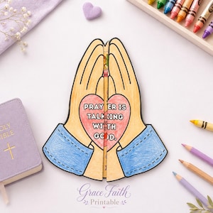 May include: A paper craft of two hands pressed together in prayer, colored with tan skin tones and blue sleeves. A pink heart in the center features the text PRAYER IS TALKING WITH GOD. The craft is split down the middle and includes a central strip.