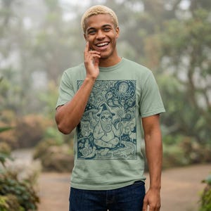Puede incluir: Camiseta verde salvia con una ilustración en azul oscuro de un perezoso mago, setas, fantasmas y otras criaturas. El diseño está enmarcado. La camiseta tiene cuello redondo y mangas cortas.
