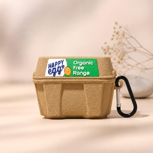 May include: A brown, egg carton-shaped case for wireless earbuds. The case has a black carabiner clip and features the text "HAPPY egg" and "Organic Free Range" on the lid. The case is made of a textured material.
