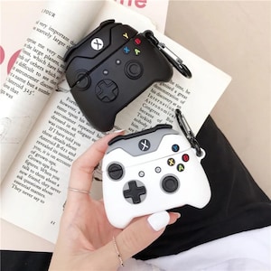 May include: Black and white AirPod cases shaped like video game controllers. The black case has a silver carabiner clip. The white case has gray accents and colorful buttons. The cases are on a book.