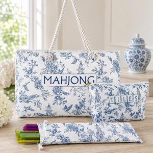 May include: A white tote bag, a matching zippered pouch, and a long rectangular case, all featuring a blue floral pattern with birds and pagodas. The tote is labeled MAHJONG, the pouch TILES, and the rope handles are white twisted cord.