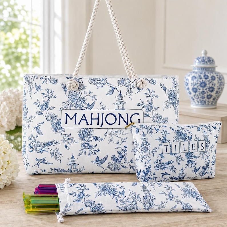May include: A white tote bag, a matching zippered pouch, and a long rectangular case, all featuring a blue floral pattern with birds and pagodas. The tote is labeled MAHJONG, the pouch TILES, and the rope handles are white twisted cord.