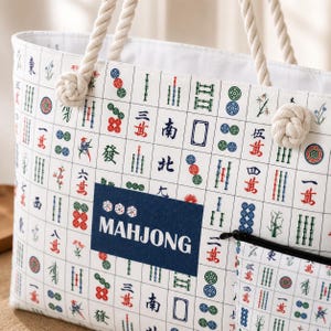 May include: A white tote bag featuring a repeating pattern of colorful mahjong tiles in red, green, and blue. The bag has thick, cream-colored rope handles and a dark blue rectangular patch in the center with three dice and the word MAHJONG.