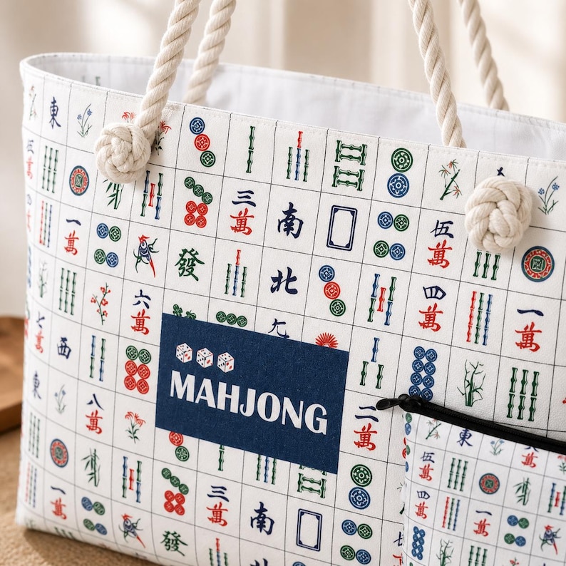 May include: A white tote bag featuring a repeating pattern of colorful mahjong tiles in red, green, and blue. The bag has thick, cream-colored rope handles and a dark blue rectangular patch in the center with three dice and the word MAHJONG.