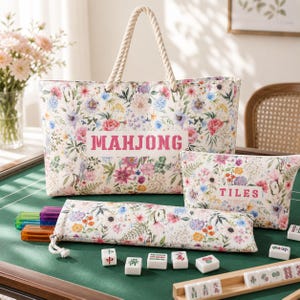 May include: A large tote bag, a smaller rectangular pouch, and a long zippered case, all featuring a colorful floral pattern on a white background. The tote displays the word MAHJONG, while the pouch features the word TILES in pink text.