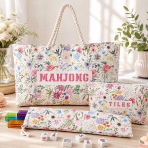 Op de afbeelding: Een draagtas met bloemenprint en touwgrepen met de tekst MAHJONG, een bijpassend tasje met rits met het label TILES, en een langwerpig etui met kleurrijke doorschijnende tegels, met enkele losse mahjongtegels verspreid op de ondergrond.