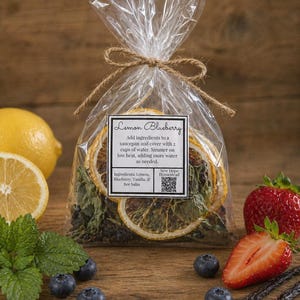 May include: A cellophane bag tied with twine, containing dried lemon slices and herbs, labelled "Lemon Blueberry." Fresh blueberries, strawberries, and vanilla pods are arranged around the bag on a wooden surface.
