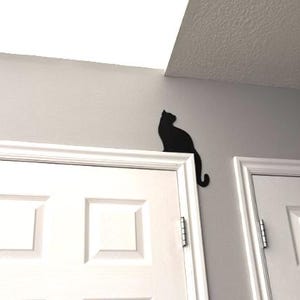 May include: A black cat silhouette wall decal positioned on the upper corner of a white door frame. The flat, solid black graphic depicts a cat sitting in profile, with its tail curving slightly downward along the gray wall surface above the door.