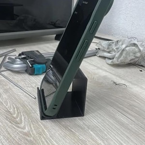 May include: A dark olive green smartphone case with a black screen, held upright by a simple black L-shaped metal stand. The case features visible side buttons and a camera bump, providing a secure fit for the device while it rests at an angle.