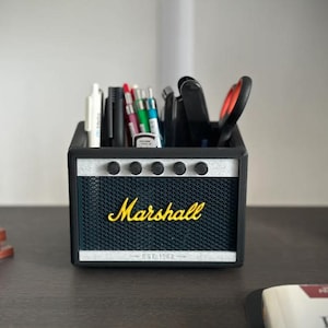 Könnte beinhalten: Schwarzer und silberner Stifthalter in Form eines Marshall-Verstärkers, gefüllt mit Stiften, Bleistiften und Scheren. Der Text "Marshall" ist in gelber Schrift. Ein kleiner Bonsai und eine Büste befinden sich im Hintergrund.
