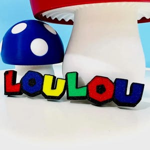 May include: A colorful, whimsical display featuring a blue mushroom with white spots, a red and white mushroom, and a vibrant, multi-colored word "LOU LOU". The letters are outlined in black.