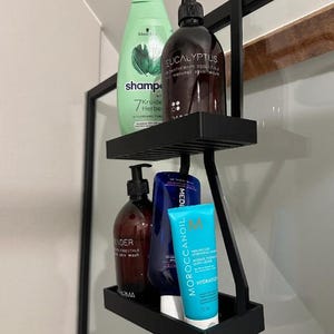 May include: A two-tier matte black metal shower caddy hanging over a glass partition. The top shelf holds a light green shampoo bottle and a dark brown pump bottle labelled Eucalyptus. The bottom shelf holds a dark brown bottle, a blue bottle, and a turquoise Moroccanoil tube.