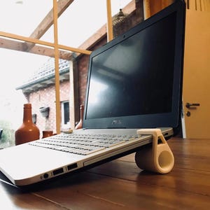 May include: An ASUS laptop in silver and black is supported by a white stand. The laptop screen is black. The stand has a cylindrical base and a curved top. The laptop is on a wooden table. The stand is made of plastic.