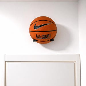 May include: An orange Nike All-Court basketball with black lines and a black swoosh logo, resting on a small black wall-mounted display bracket. The ball features the text ALL-COURT INDOOR/OUTDOOR printed in black on its textured rubber surface.