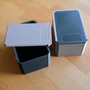 以下が含まれることがあります： グレーと濃いグレーの3次元長方形収納ボックス。1つのボックスが開いており、黒い内部が見えます。蓋にはリブのデザインがあります。ボックスは明るい茶色の木製の表面にあります。