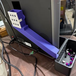 May include: A purple 3D-printed chute attachment mounted to the back of a computer monitor, featuring a white rectangular top section with a grid of nine holes. A black rectangular bin sits below the chute, containing small purple and white plastic scraps.
