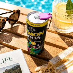 May include: A black aluminum can of Credo Lemon Soda with yellow text and a graphic illustration, featuring a pink plastic tab opener attached to the top with a small metal ball chain, positioned next to sunglasses, a glass, and a magazine.