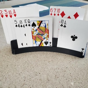 May include: A black playing card holder displaying a hand of cards. The cards show various suits and values, including hearts, diamonds, clubs, and spades. The holder is on a speckled countertop.