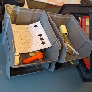 May include: Two grey plastic storage bins with open fronts. The left bin contains a white paper sheet with a grid of flower icons and an orange plastic tool. The right bin holds various metal screws, a yellow plastic piece, and a small white rod.