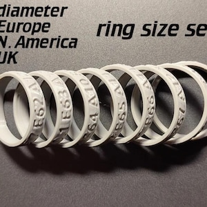 May include: A set of seven light gray ring sizers arranged in a row. Each ring features embossed alphanumeric codes including E62, E63, A10, B65, and B67. The text "diameter Europe N. America UK ring size sets" is printed in black above the rings.