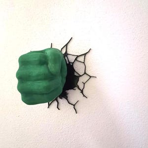 May include: A three-dimensional wall-mounted sculpture featuring a green fist emerging from a black, jagged crack design. The fist is textured with defined knuckles and fingers, creating the appearance of breaking through the wall surface.