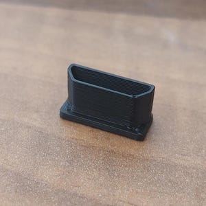 Puede incluir: Un pequeño componente negro de forma rectangular impreso en 3D, con una abertura superior hueca y cónica y una base plana más ancha. El objeto muestra líneas de capa horizontales visibles a lo largo de sus paredes exteriores.