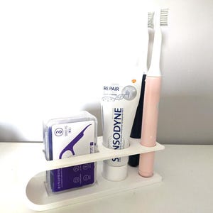 May include: A white plastic bathroom organiser stand featuring three circular cutouts. It holds a purple box of dental floss picks, a tube of Sensodyne Repair toothpaste, and a light pink electric toothbrush with a white brush head.