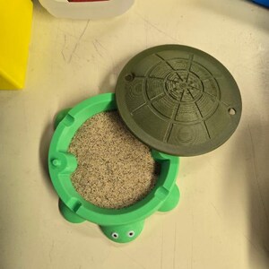 May include: A bright green turtle-shaped container with googly eyes, filled with light tan sand. The container features a removable dark olive green circular lid with a textured, radial pattern on its surface, resting partially off the base.
