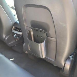 May include: A black, rectangular storage bin attached to the back of a car seat. The bin is made of a rigid material and has a simple, open design. It is mounted with two screws and is designed to hold small items.