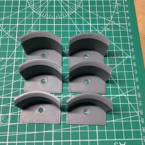 May include: Six grey, 3D-printed, semi-circular mounting brackets arranged in two columns of three. Each bracket features a curved support surface and a flat base with a single centered circular hole for mounting, resting on a green cutting mat.