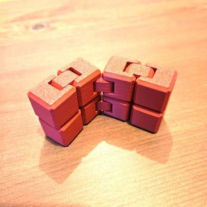May include: A red fidget toy constructed from a series of interconnected, articulated cubes. The object features a matte, textured surface with visible 3D printed layer lines, arranged in a flexible, hinged chain that allows for various folding configurations.