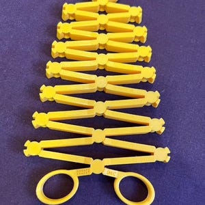 Könnte beinhalten: Ein ausziehbares Scherwerkzeug aus gelbem Kunststoff mit einem gitterartigen Klappmechanismus. Die Basis umfasst zwei Fingerringe mit dem eingeprägten Text SCISSOR und SHAPE am Griff. Das obere Ende endet in einer kleinen, gezackten Greifklaue.