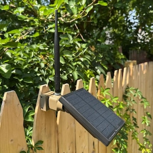 May include: A tan-coloured mounting bracket attached to a wooden fence, featuring a vertical black antenna and a rectangular black solar panel angled upwards to capture sunlight, designed for outdoor electronic equipment power and connectivity.