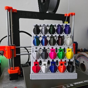 May include: A 3D printer with an orange frame and a display of small, colorful 3D-printed objects. The objects are arranged in a grid, showcasing a variety of colors including red, blue, green, and yellow. A spool of filament is visible.