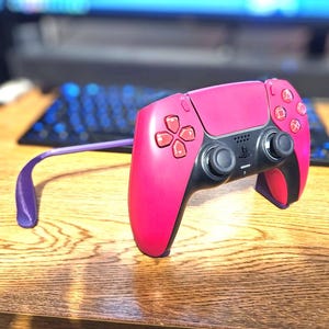 May include: A bright pink wireless video game controller featuring black accents on the central panel and analogue sticks, with pink directional buttons and action symbols. A purple plastic arm extends from the side of the controller body.