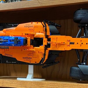 May include: A top-down view of a bright orange and blue Formula 1 race car model. The car features black tires, a white support stand, and various sponsor logos including Webex, Darktrace, and Dell printed on the bodywork and front wing.