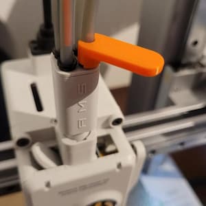May include: A white 3D printer filament buffer component labelled AMS with a bright orange plastic lever attached to the top. Four translucent filament tubes are inserted into the top of the white housing, which is mounted on a mechanical frame.