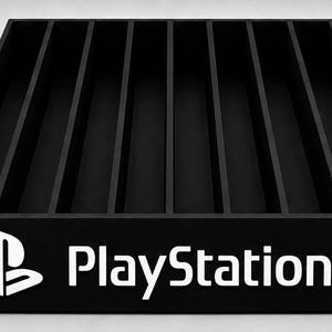 Może przedstawiać: Czarny, prostokątny organizer do przechowywania z ośmioma pionowymi przegródkami na pudełka z grami. Na przednim panelu znajduje się białe logo PlayStation oraz napis PlayStation wykonany bezszeryfową czcionką, co zapewnia uporządkowaną ekspozycję.