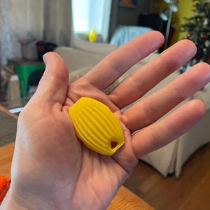 May include: A small, bright yellow, oval-shaped object with a ribbed, textured surface and a single hole at one end. The object features a symmetrical design with vertical ridges running along its length, providing a distinct tactile pattern.