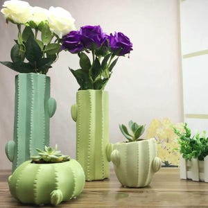 Puede incluir: Dos jarrones cilíndricos altos con forma de cactus en verde claro y verde menta, con rosas artificiales blancas y moradas. Dos macetas redondas más pequeñas con forma de cactus en verde claro contienen suculentas, junto a una caja de madera blanca.