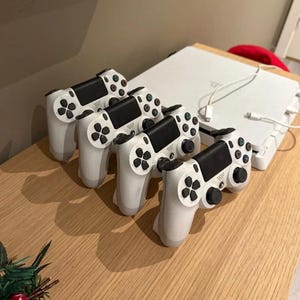 May include: Four white wireless game controllers with black touchpads, joysticks, and buttons arranged in a row next to a white gaming console with a connected charging cable. The controllers feature the standard directional pad and action buttons.