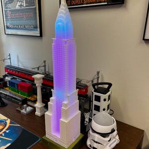 May include: A white 3D-printed model of the Chrysler Building featuring a stepped, tiered design with a pointed spire. The structure is illuminated from within by blue and purple LED lights, creating a glowing effect throughout the skyscraper model.