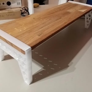 May include: A wooden monitor stand with a light brown surface and white legs. The stand is rectangular and has a modern design. The legs have a geometric pattern.